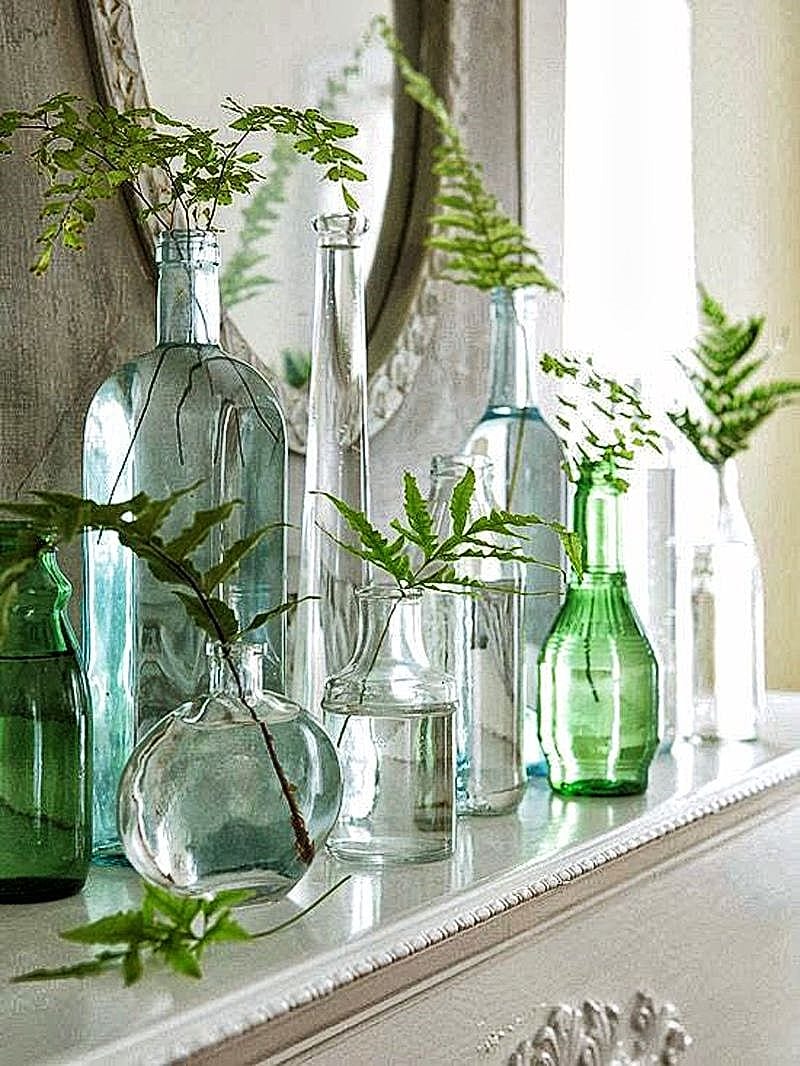 recycled green and clear glass bottles of various sizes and heights and fern branches with circular mantel mirror and white mantel with dentil details