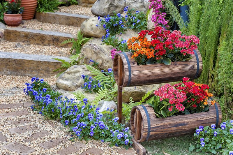 small barrels with cutouts to accommodate flowering plants and barrel stands with wooden stairs with pea gravel steps and pansies on the ground and in between boulders