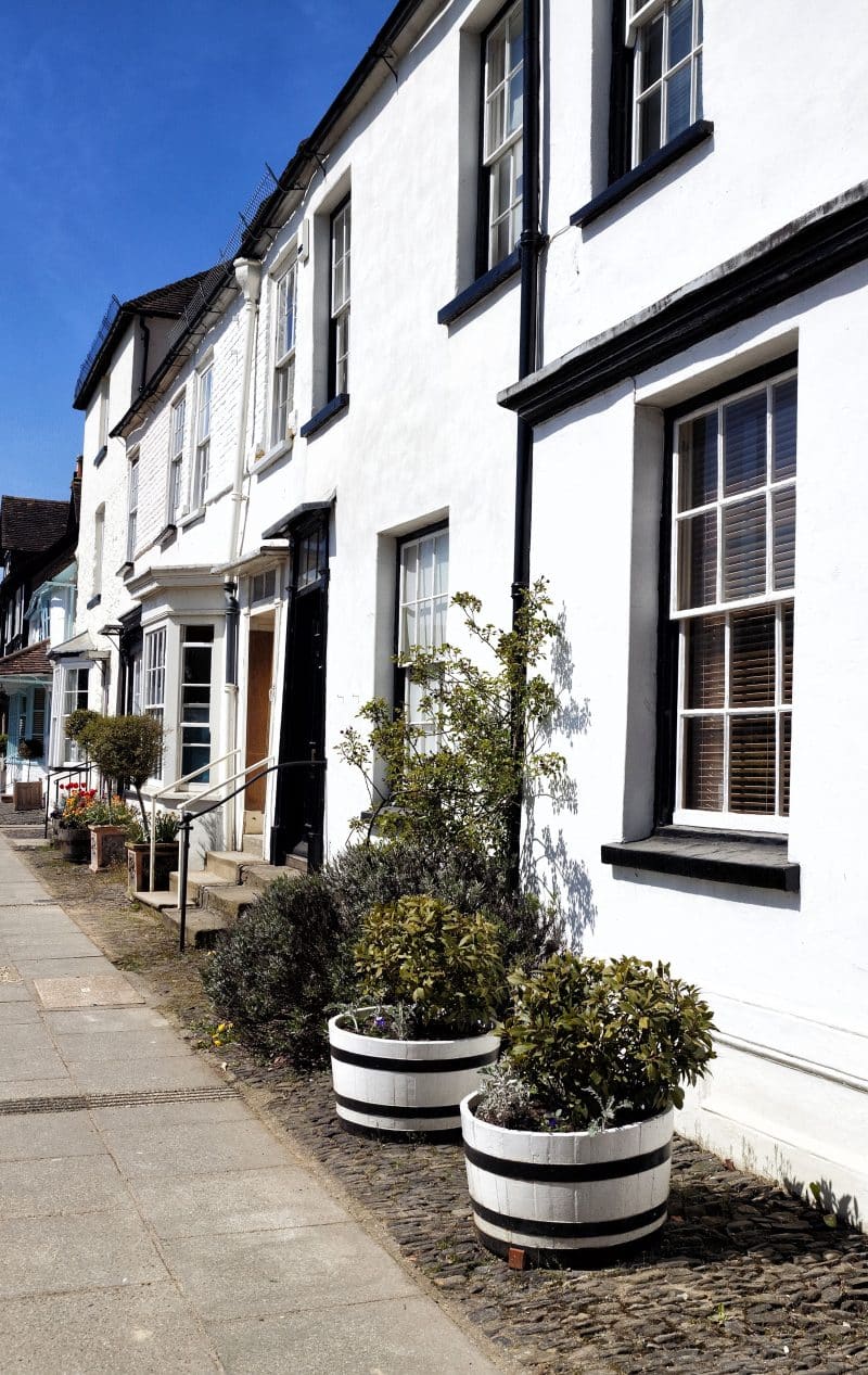 white half barrels on one side and twin aged barrel planters on the other side with contrasting black barrel hoops and square engraved terra cotta tree planters and painted wooden window and door canopies with bay windows