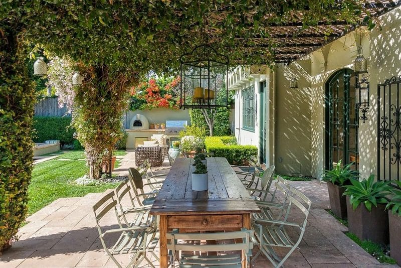 lattice pergola with climbing vines with oversized hanging lantern candle holder over farmhouse table and pale sage green folding metal bistro chairs with segmental arched pine green French doors and 3 tapered square old stone planters