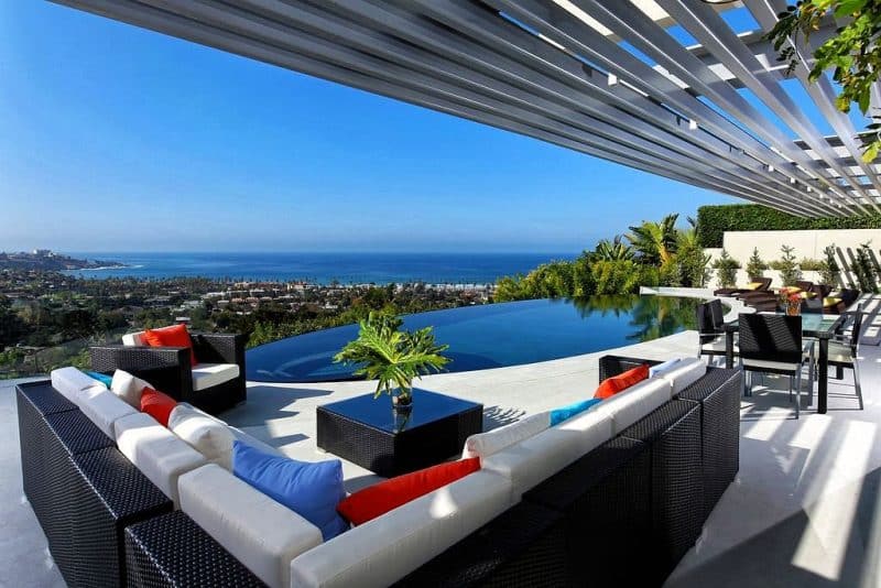 long silver gray aluminum pergola with oval infinity pool and black L-shaped wicker sofa with mixed throw pillows and glass-topped square wicker coffee table with split-leaf philodendron in hurricane vase