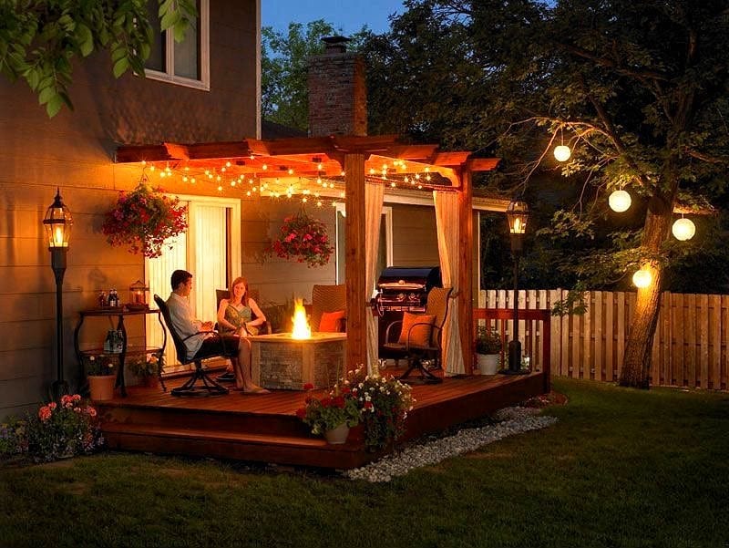 red cedar pergola with string lights and 4 resin wicker swivel chairs with round base and square faux stone propane fire pit table with stone veneer exterior walls and 2 black powder coated outdoor gas lamps