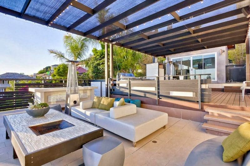 steel attached pergola with mesh shade and smaller rafters in between with string lights on pergola and stainless steel pyramid flame outdoor gas heater with linear mosaic tiled fire pit table and ecru modern modular sofas with 2 light gray sculptural garden stools