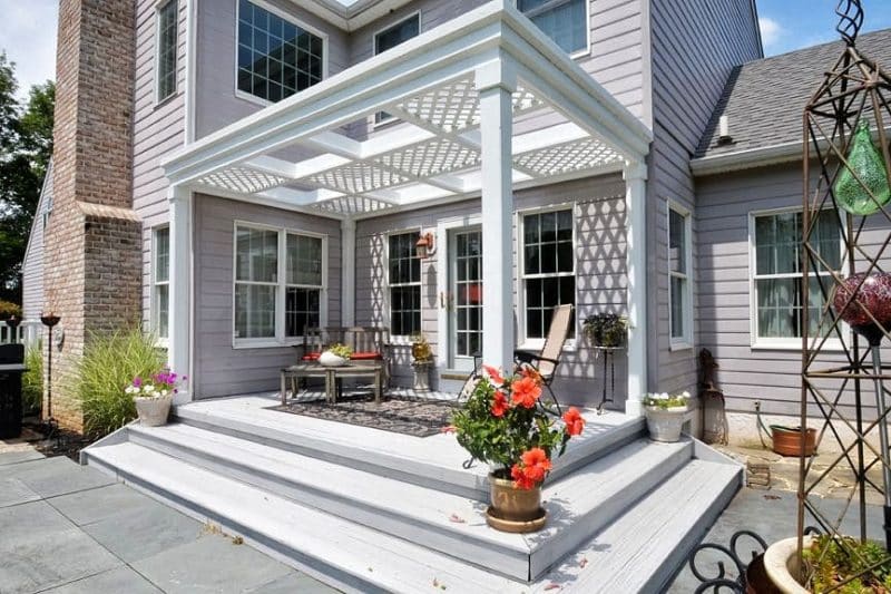 white attached pergola with combination open-top and lattice panels on glass storm door with copper handle matching the outdoor wall sconce and petunias and hibiscus flowering plants on patio stairs