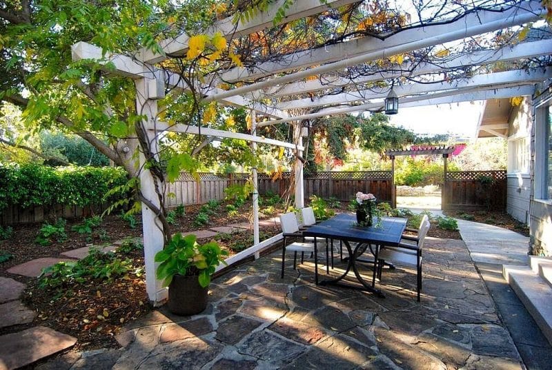 white pitched pergola attached to clapboard wall and small lantern pendant light over dining area with board and batten fence with lattice top panel and entry arbor with flagstone pathway with matching pergola floor