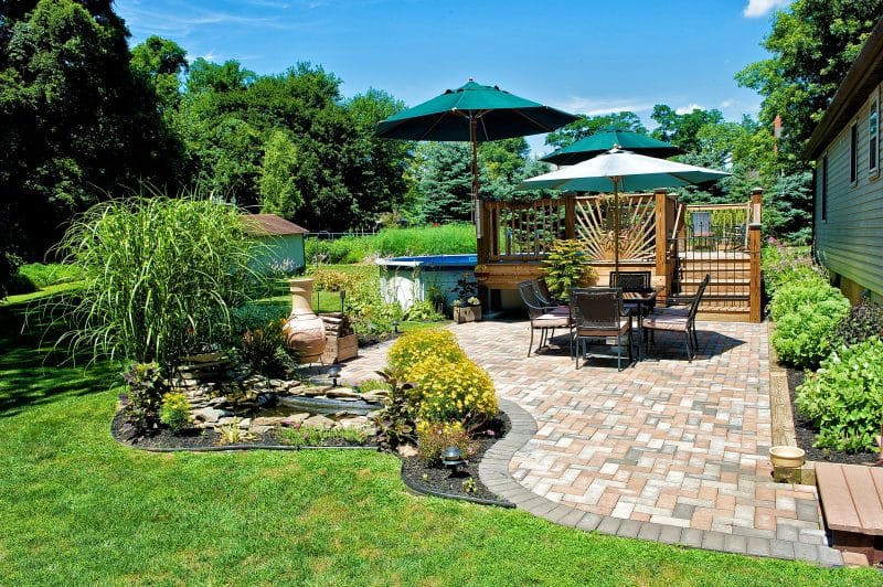 curved soldier course brick patio edging with small pond with flat fieldstone edging and resin black landscape edging with timber edging on slightly raised bed and above ground pool deck with 2 patio umbrellas attached to deck balustrades with decorative deck railing