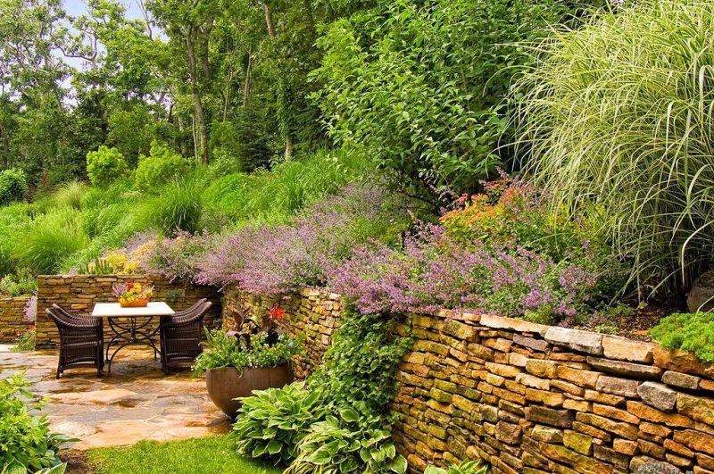 dry stacked stone patio fence with Mexican bush sage and 4 chocolate brown outdoor wicker armchairs with marble topped table with curved corners and legs and large stained concrete bowl planter with mixed plants