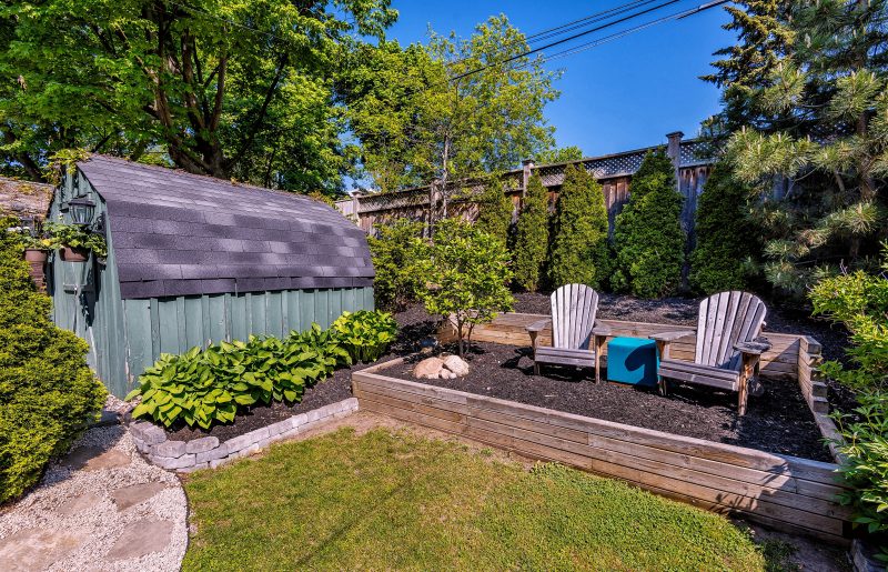 gray brick-shaped stone edging and large raised bed with wood retaining walls and high back side with 2 Adirondack chairs on raised bed with dark turquoise square side table and small sage green garden shed with gray felt roof