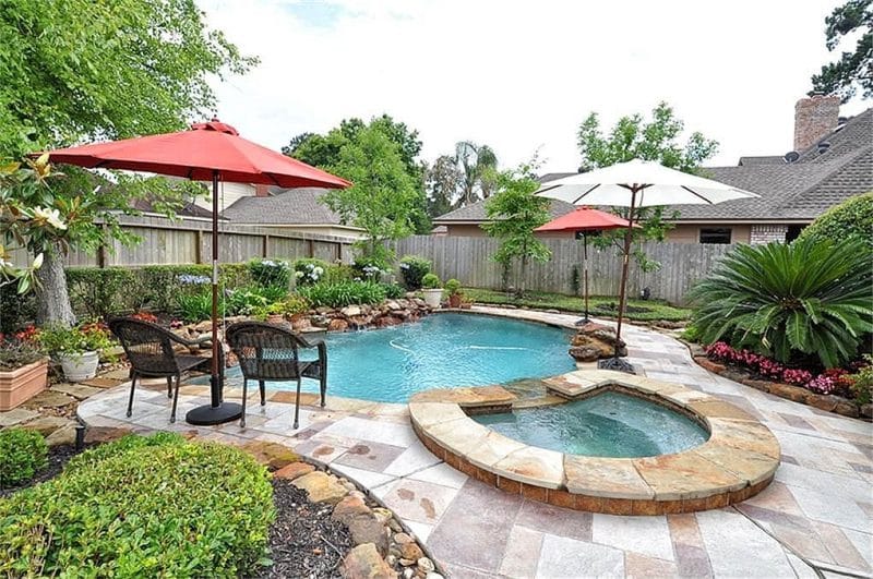 freeform shaped swimming pool with rounded spa and Agapanthus midnight blue plants on boulder style waterfall and rock edged garden beds bordering travertine pool decking with 2 red and 1 white large deck umbrellas with 2 resin wicker armchairs under red deck umbrella
