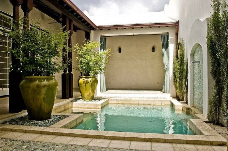 geometric spool pool with steps and oil-rubbed bronze Roman scupper on arched and tiled wall niche with 2 large olive outdoor ceramic planter flanking pool steps and travertine coping and pebble deck tiles with poolside shower and changing area with green outdoor drapery