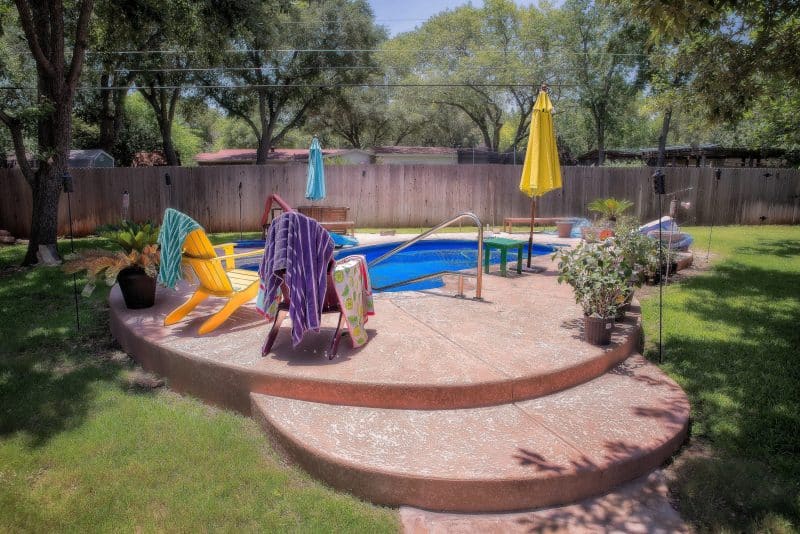 kidney-shaped swimming pool with polished steel deck mounted stair rails and towels draped on painted Adirondack chairs and dog ear wooden panel fence with blue and yellow closed poolside umbrellas and concrete pool decking with stained acrylic surface
