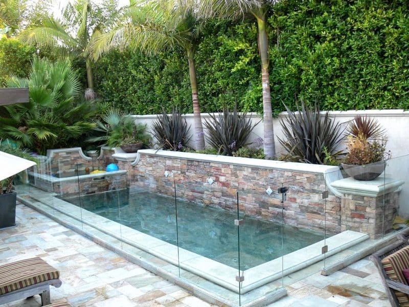 narrow rectangular lap pool with raised spa and frameless glass pool fence with travertine tiles on swimming pool deck and raised stone veneer pool wall with water feature and 2 low bowl planters on capped stone pedestals