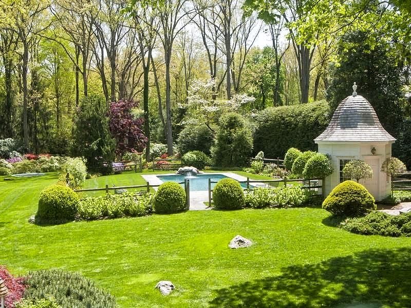 stone waterfall on one end of pool and pool house with bell-shaped roof and Western style rail log fence and artificial turf surrounding swimming pool area with topiaries accenting border hedges