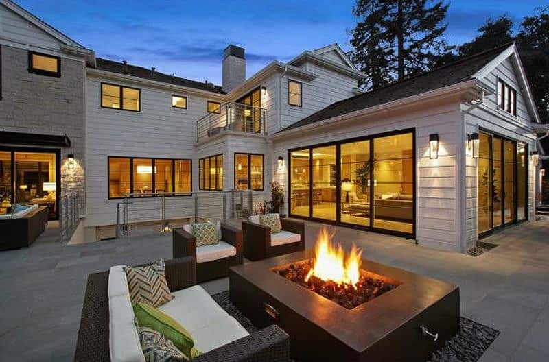 brown concrete fire pit table on river rocks and green and patterned throw pillows on white sofa cushions with stainless steel handrails with cable railing and white shiplap wall cladding with outdoor light sconces with soft lighting seen through windows and glass patio doors