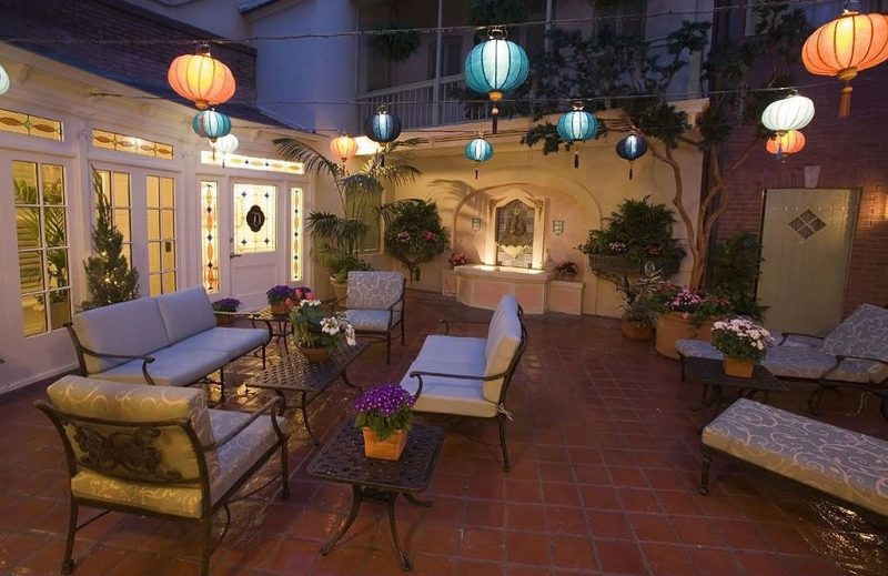 multicolored lantern string lights with tassels and stained glass panels on white framed door, sidelights, and transoms with various sized terracotta planters with flowering plants on side tables and wrought iron patio seats and chaise loungers with jacquard vine pattern brocade fabric cushions with arched wall fountain with lighted catch basin on stucco wall