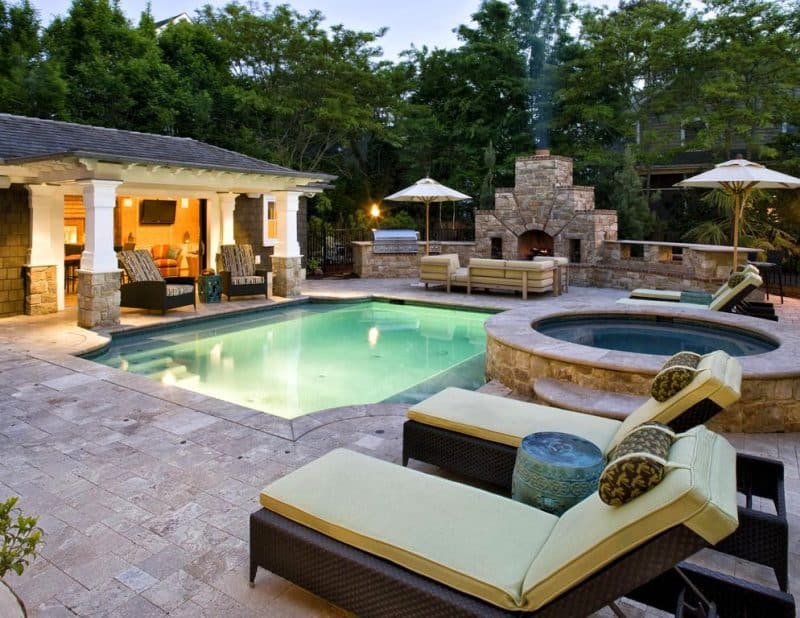 Grecian shaped swimming pool with underwater LED lights and outdoor kitchen with grill and sectional sofa on seating area with ceramic drum-shaped side tables between outdoor wicker adjustable chaise lounges with custom contour headrest pillows and tiered outdoor wood-burning stone fireplace with built-in firewood niches and poolside patio covered pergola with stone veneer base column wraps